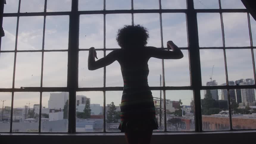 A young woman dances next to a glass window with a city view visible through the window at sunset