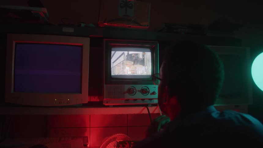 A person is sitting in a dim room with red lighting looking at three screens the central screen is a small old television displaying footage of a construction site the other two screens appear to be older computer monitors