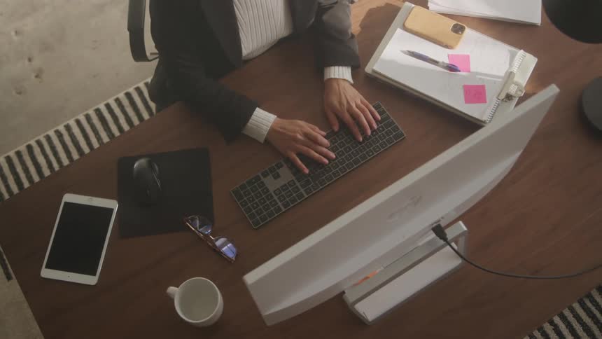 A young woman in her office wearing a suit uses her phone then drops it and begins typing on her computer