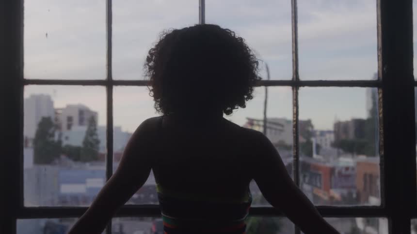 A silhouetted person with curly hair stands in front of a large gridded window stretching their arms out as they look at the blurry cityscape outside