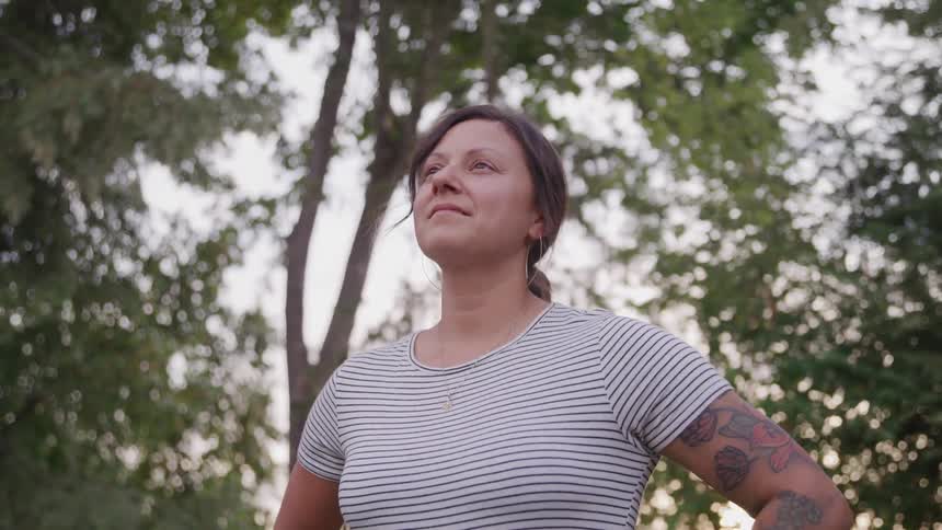 A woman with dark hair tied back is standing outdoors looking up and to the side with a determined expression she is wearing a striped tshirt and has tattoos on her arm with a backdrop of green trees