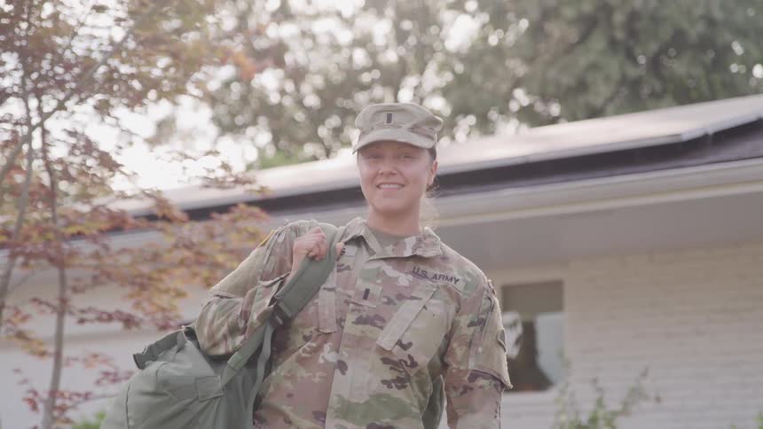 A young female soldier in a camouflage uniform and hat smiles while carrying a duffel bag over her shoulder in front of a house