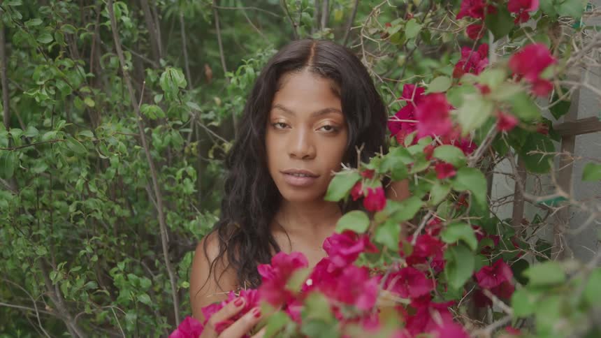 A woman stands among lush greenery and flowers looking directly at the camera
