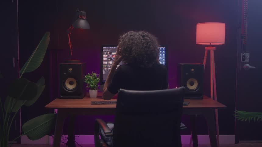 A creative professional with curly hair sitting in an office chair at a desk stands up and walks away from their computer monitor which is displaying a video editing timeline