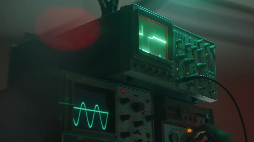 A mans hands are shown turning knobs on a complex piece of electronic equipment a green waveform is displayed on a screen with another screen below showing a sine wave