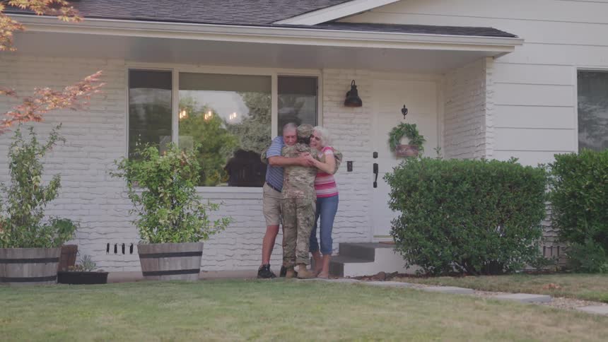 A woman wearing a military uniform runs and hugs her elderly parents standing on their porch at midday all looking excited