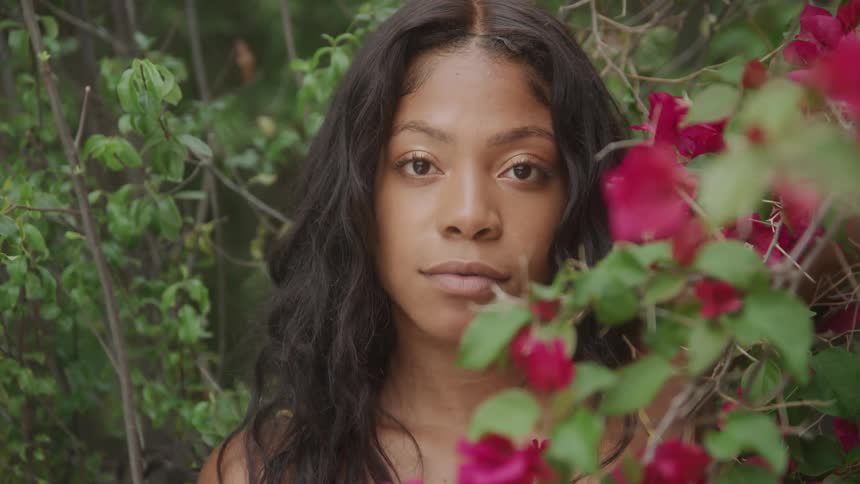 A serene woman with long dark wavy hair looks up at the camera while standing among green leaves and vibrant pink flowers
