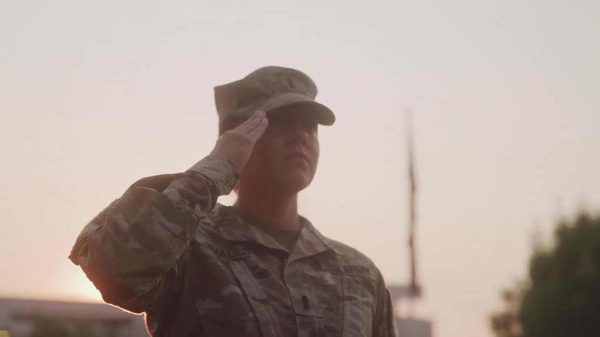 A military officer stands outdoors in a salute pose with the setting sun visible