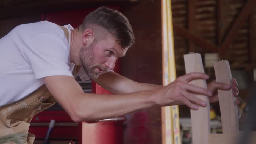 A bearded man in a white tshirt and a brown apron is working on a piece of lightcolored wood in a workshop he is intently focused on his work and using his hands to guide the wood