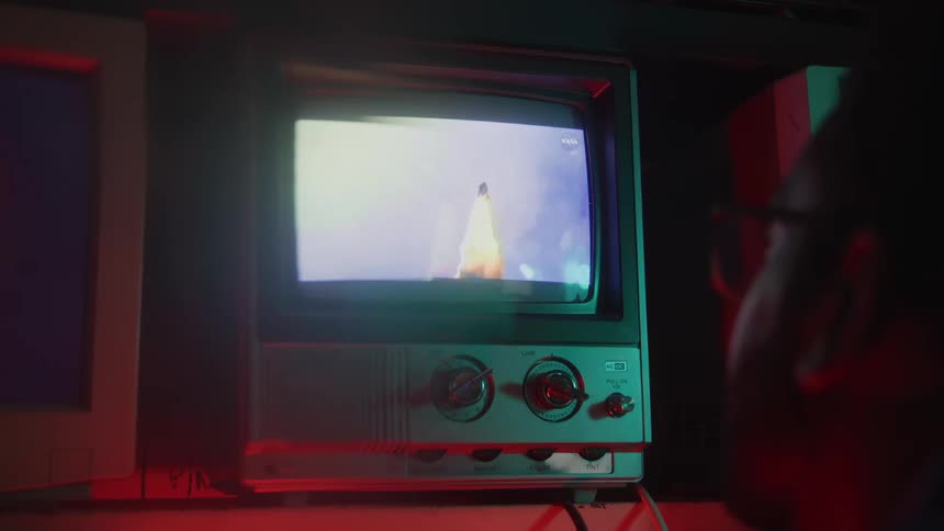 A retro television displays a rocket launch with a large cloud of smoke and fire at its base a persons head seen from the back is visible in the foreground as they watch the screen