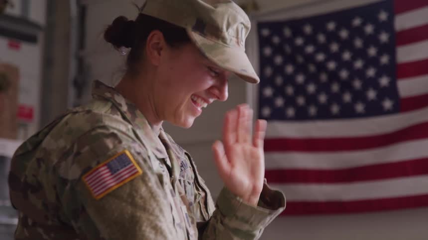 A happy female soldier in a camouflage uniform with an american flag patch on her shoulder waves and smiles in front of a large american flag