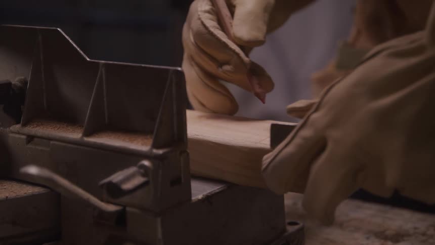 A carpenter wearing thick tan leather work gloves places a block of wood into a miter box to prepare it for cutting