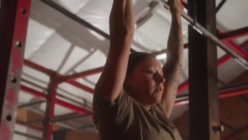 A female athlete in a tan tshirt with tattooed arms claps her hands together creating a cloud of chalk dust before doing pull ups in a dimly lit gym