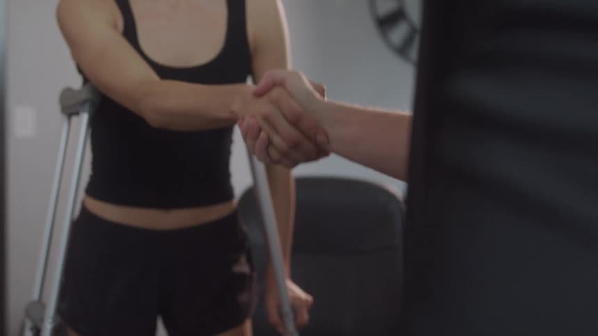 A male physical therapist in a black shirt shakes hands with a female patient wearing a black tank top and shorts who is standing with crutches
