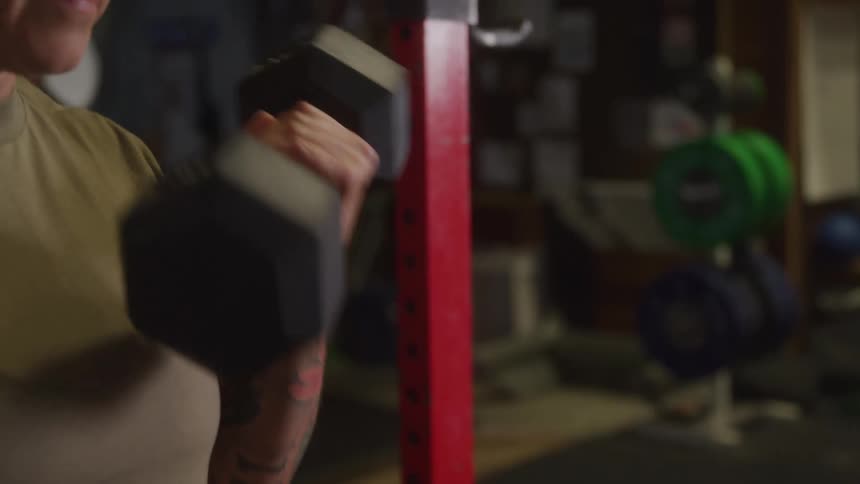 A tattooed woman wearing a tan tshirt and dark pants curls heavy black dumbbells in a gym