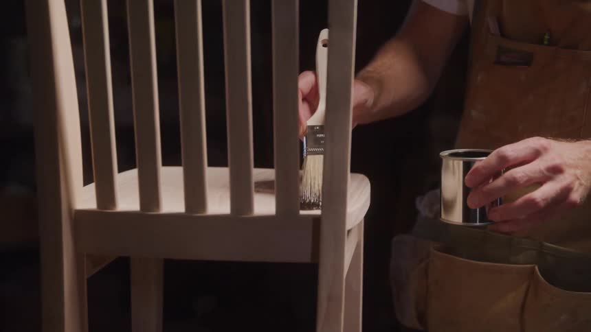 A worker in a workshop applies a clear coat to a wooden chair with a paintbrush the chair appears to be lightcolored and the clear coat makes the wood a slightly richer darker color