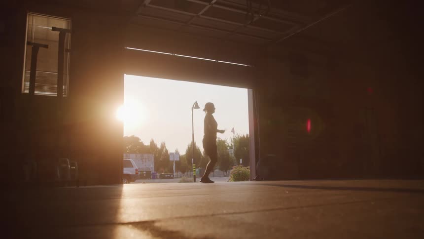 A silhouette of a person jumping rope in a garage or warehouse is seen from a low angle the person is in a welllit doorway with a suburban street and trees visible in the background