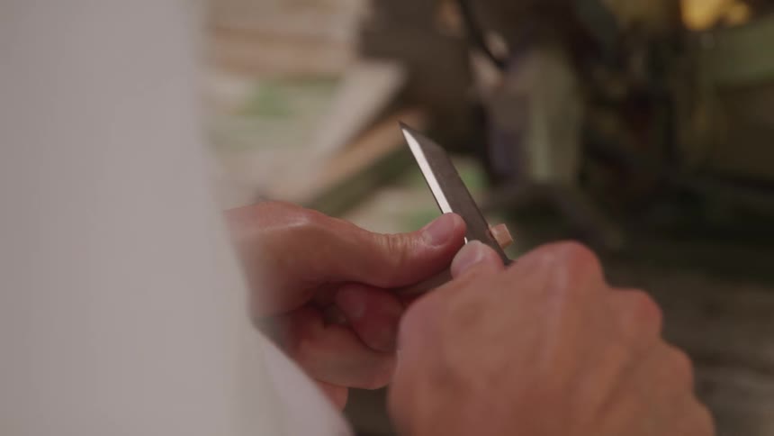 A woodcarver wearing a white shirt holds a small block of wood and uses a sharp knife to carefully whittle a precise curve into it