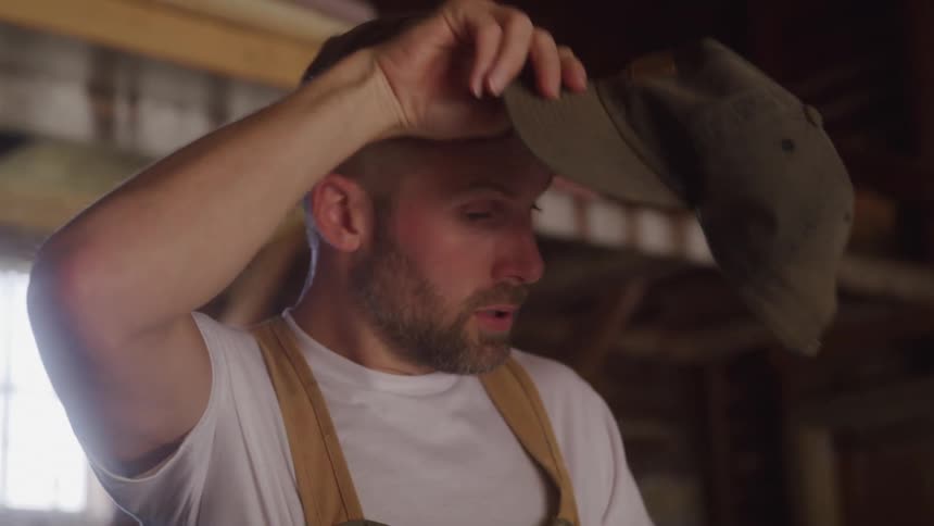 A man holding a broom takes off his hat to wipe the sweat from his face then puts it back on and resumes sweeping