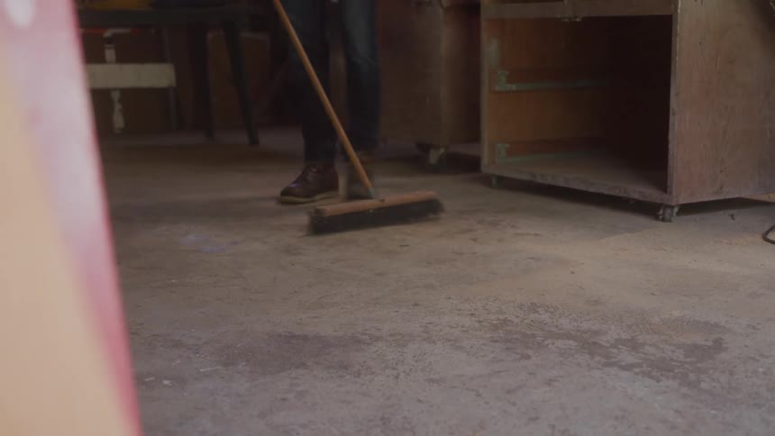 A person wearing jeans and brown boots sweeps the concrete floor of a workshop with a wooden handled push broom