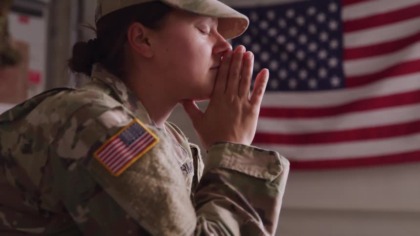 Uniformed woman in the us military closes her eyes and clasps her hands in a reflective gesture in front of the american flag