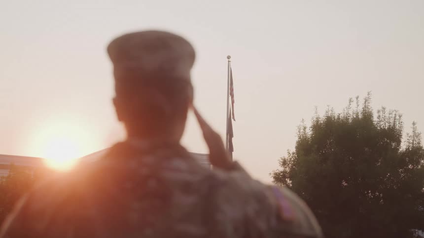 A blurred figure in a military uniform is seen from behind facing away from the camera they raise their hand in a salute toward a flagpole with a blurred flag waving in the distance against the backdrop of a building and a setting sun