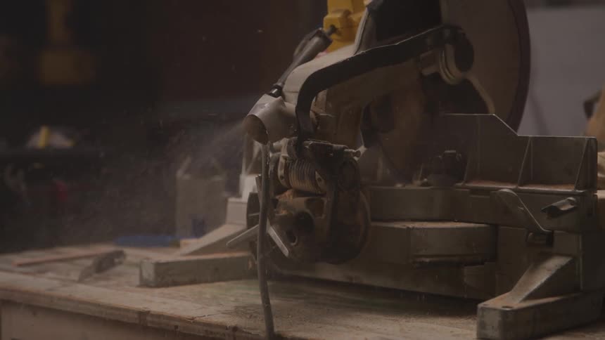A stationary miter saw on a workbench has a cloud of wood dust and debris blowing away from it while cutting