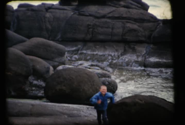 A kid running on some huge boulders and jumping down on the beach and running towards the camera