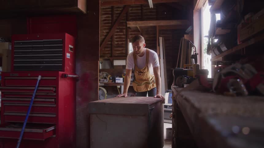 Man in an apron rests his hands on a workbench inside a workshop