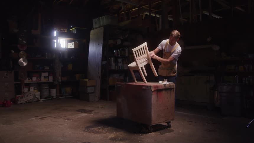 Man works on a wooden chair placed on a block in a dimly lit workshop