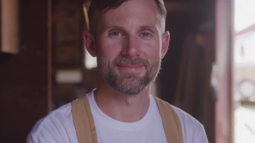 Man with a pencil behind his ear smiles slightly inside a workshop