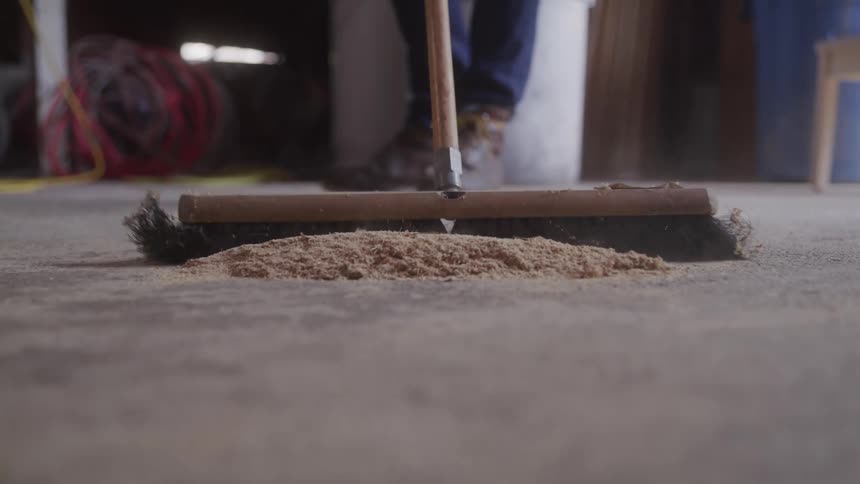 A wide push broom sweeps a pile of wood shavings and dust across a concrete floor in a workshop