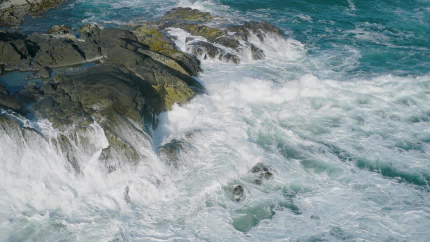 Ocean waves crash forcefully against a rocky coastline creating a dramatic spray of white foam the water is a deep bluegreen contrasting with the dark gray rocks