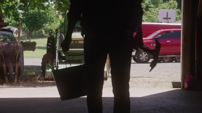 A silhouette of a farmer carrying a bucket and a shovel stands in the doorway of a barn or garage with a green tractor and a red truck visible outside on a sunny day