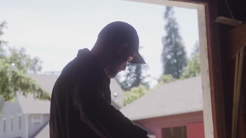 A silhouetted person wearing a baseball cap stands inside a garage or warehouse with a bright doorway behind them the person is holding a long thick rope and appears to be working with it
