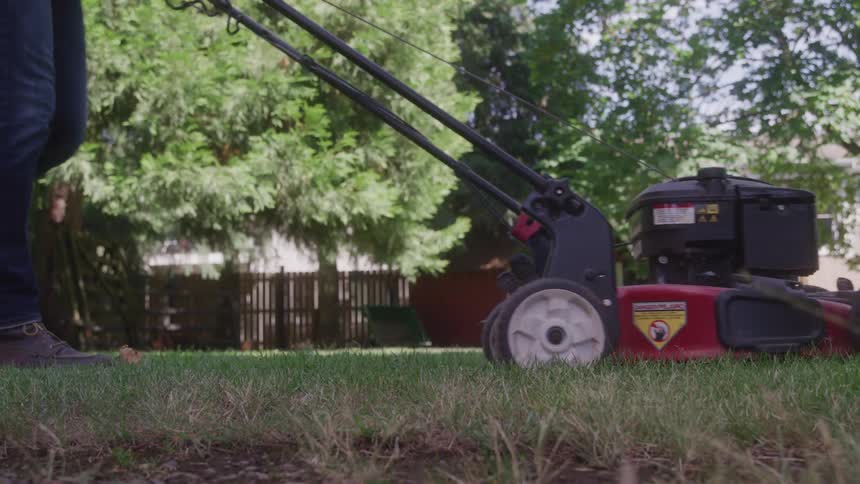 A low angle view shows a person wearing jeans and brown shoes pushing a red and black lawnmower across a lawn