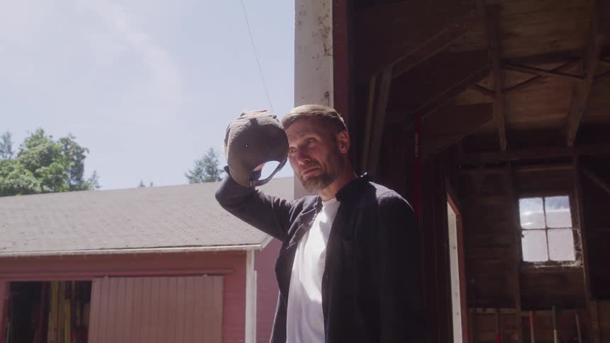 A man wearing a baseball cap raises his hand to shield his eyes from the sun as he stands in a doorway of what appears to be a barn or shed the background shows a bright sunny day with trees and a building visible outside