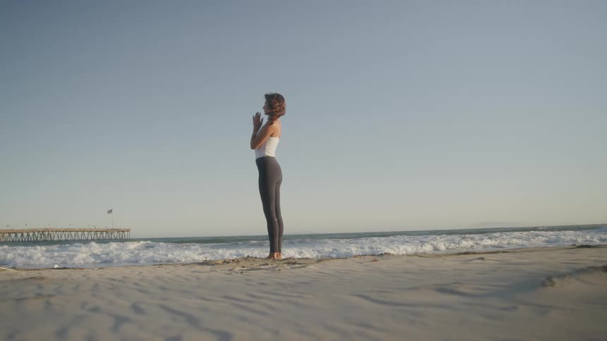 Woman standing on the sand holds a prayer pose facing the ocean