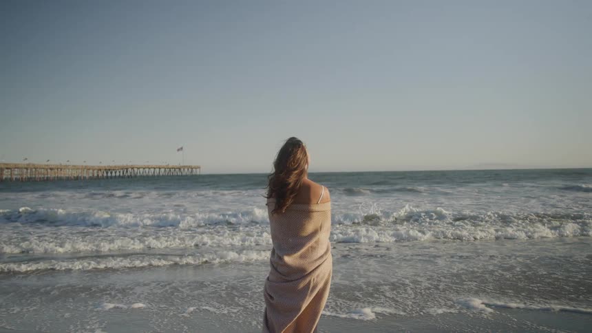 Woman wrapped in a blanket watches the sea near a pier in the late afternoon