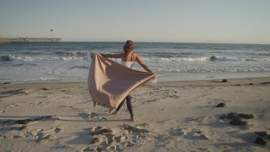 A woman walks barefoot on the beach at midday holding a blanket and having fun