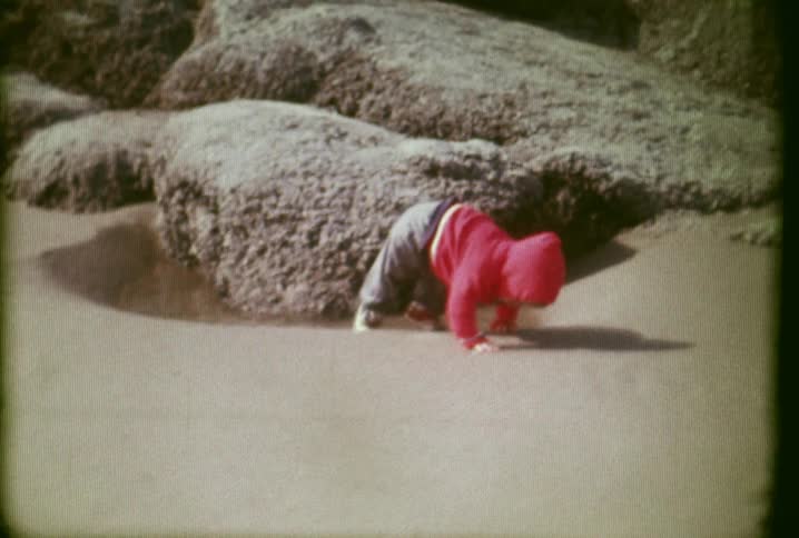 A child in a red jacket crawls out of a puddle on the beach