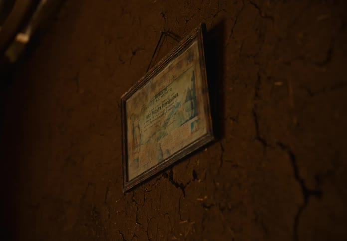 A framed certificate hangs on a cracked earthen wall in dim light