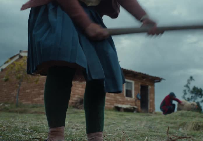 A woman tills the soil with a hoe while another person pets a sheep with a hut in the background on a cloudy day