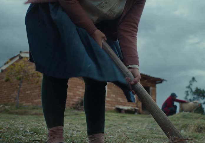 A woman tills the soil with a hoe while another person pets a sheep with a hut in the background on a cloudy day