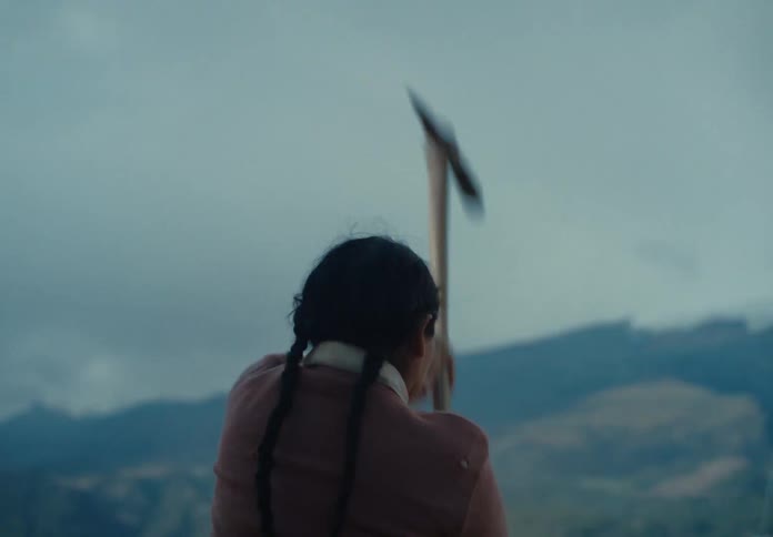 Woman with a braid works with a hoe in a mountainous region under a cloudy sky