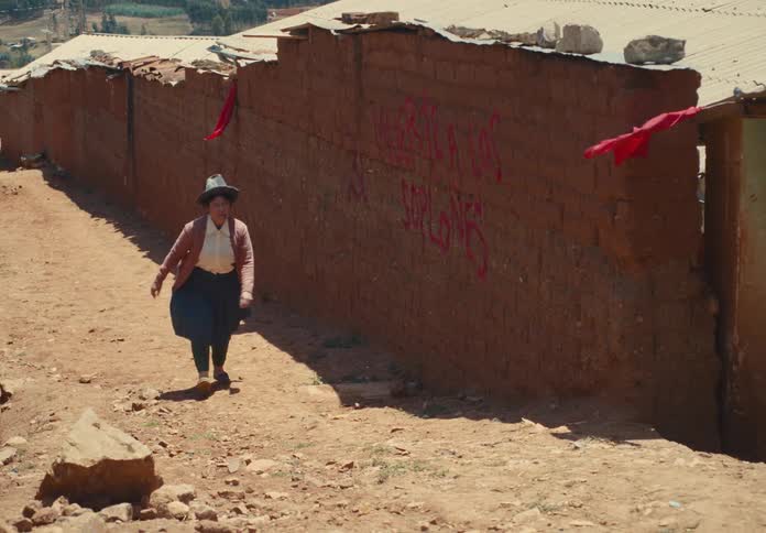 Woman walks along a dirt road beside adobe buildings with red flags hanging
