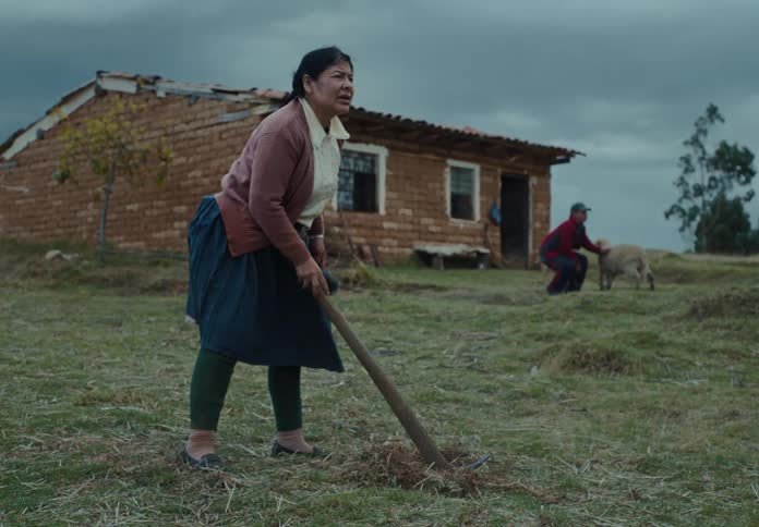 Woman works the land with a hoe in front of a mud brick house and a man in the background with a calf
