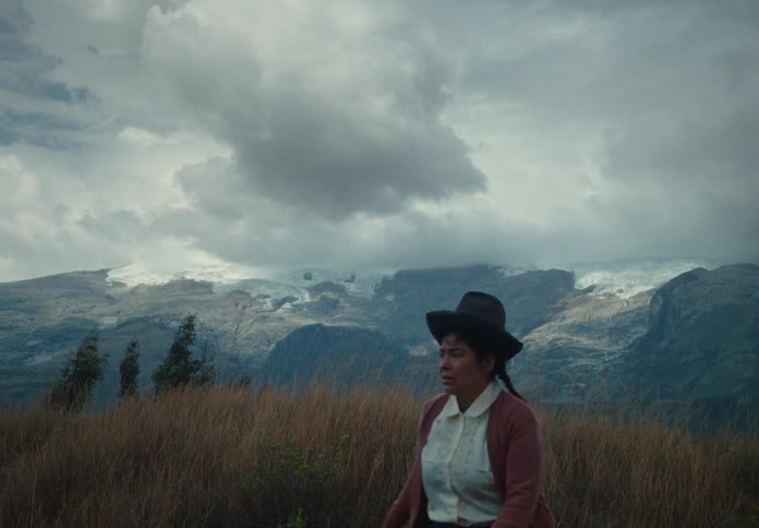 Woman walks through open field with mountains in the background under a cloudy sky