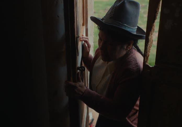 A female peers cautiously through a narrow opening in a dark wooden door and opens the door