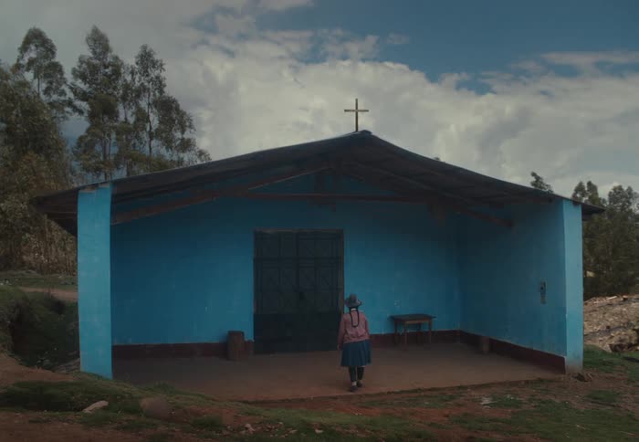 Woman walks toward a small blue church with a cross on the roof surrounded by trees and a partly cloudy sky